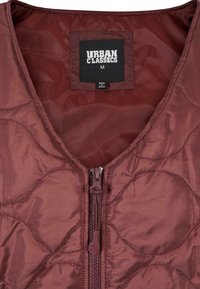 Mörkröd kviltad väst med dragkedja framtill och rundad halsringning. Inkluderar en etikett med "Urban Classics" och storlek "M." Slät, glänsande material.