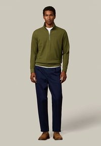 Pull zippé vert olive avec col montant, associé à un pantalon bleu marine. La tenue présente une coupe classique et un tissu texturé.