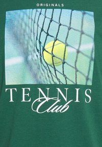 Grön t-shirt med ett fotografiskt tryck av en tennisboll nära ett nät, med texten "ORIGINALS" och "TENNIS Club" i vitt.