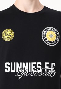 T-shirt en coton noir avec deux logos circulaires aux accents jaunes et un texte blanc qui dit "SUNNIES FC Lyle & Scott." Texture lisse.