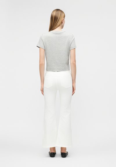 Γυναίκα με μακριά ξανθά μαλλιά που φοράει γκρι κοντομάνικο τοπ, λευκό παντελόνι με βολάν και μαύρα παπούτσια, στέκεται με την πλάτη στραμμένη προς τα εμπρός.