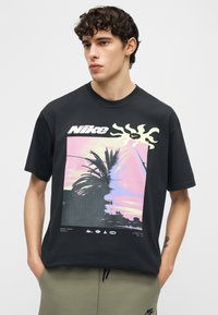 Νεαρός άνδρας με σγουρά μαλλιά που φοράει μαύρο t-shirt Nike με πολύχρωμο σχέδιο φοίνικα και ηλιοβασίλεμα, στέκεται μπροστά σε απλό φόντο.