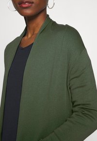 Grön stickad cardigan med öppen framsida, texturerad tyg, långa ärmar och en mjuk, avslappnad passform, bärs över en mörk v-ringad topp.