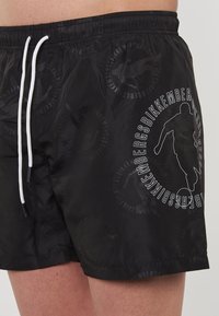 Pantaloncini da bagno neri con una texture a motivo, coulisse bianca a contrasto e un logo stampato con design a silhouette.