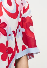 Κοντινό πλάνο κοντού μανικιού με κόκκινο, λευκό και ανοιχτό μπλε αφηρημένο λουλουδάτο και γεωμετρικό σχέδιο σε ελαφρύ ύφασμα.