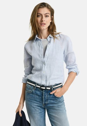 Lichtblauw linnen shirt met een knoopsluiting aan de voorkant, opgerolde mouwen, in combinatie met vervaagde blauwe jeans en een zwart-wit gestreepte riem.