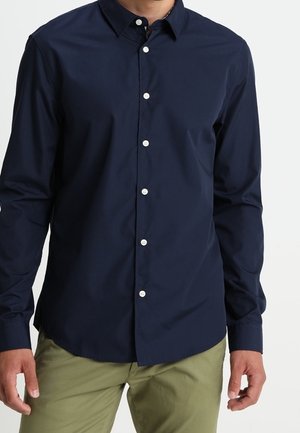 Homme portant une chemise bleu marine à manches longues boutonnée et un pantalon vert olive, debout les bras détendus le long du corps.