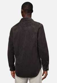 Mężczyzna w ciemno-brązowej koszuli z korduroy z długim rękawem, pokazany od tyłu, w zestawieniu z jasnobeżowymi spodniami na jednolitym tle.