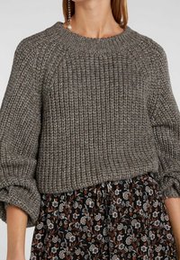 Gruby szary sweter dziergany o prążkowanej fakturze, z okrągłym dekoltem i obszernymi rękawami, zestawiony z czarną spódnicą w kwiaty z pomarańczowymi akcentami.