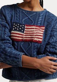 Blauer Pullover mit Zopfmuster und einer aufgestickten amerikanischen Flagge. Gerippter Kragen und Bündchen; durchgehend strukturiertes Muster.