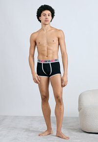 Chiloți boxer negri cu talie elastică roșie și albă, cu brand. Materialul pare elastic, cu o textură netedă. Modelul stă pe o suprafață texturată.