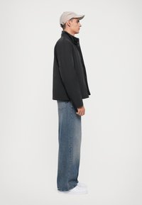 Svart, knappad jacka med hög krage, bärs över lösa blå jeans och vita sneakers. Ljusbeige keps fulländar outfiten.
