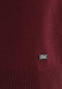 Bordeaux gebreide stof met geribbeldeZoom en een klein zwart metalen label met de tekst "Petrol Ind" dat dichtbij de onderrand is genaaid.
