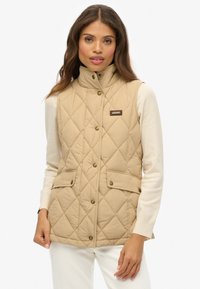 Gilet matelassé beige avec un col haut, des boutons-pression et des poches avant. Porté par-dessus un haut à manches longues de couleur claire, avec des éléments de design minimalistes.