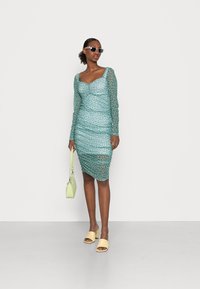 Vestido de malha turquesa com bolinhas, mangas longas e design franzido. Combinado com mules de salto bloco amarelos e uma pequena mala verde.