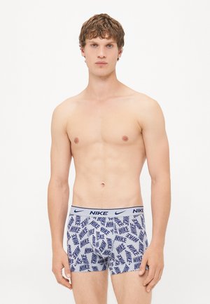 Junger Mann mit heller Haut und lockigem Haar, der leichte graue Nike-Boxershorts mit navyblauem Logo-Muster trägt, steht vor einem weißen Hintergrund.