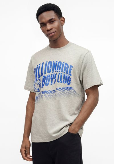 Νέος άνδρας που φορά γκρι ανοιχτό T-shirt με μπλε γράμματα "Billionaire Boys Club" και λογότυπο αστροναύτη, στέκεται με το ένα χέρι στην τσέπη.