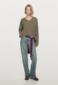 Olivgrön kabelstickad tröja med V-hals, ihopparad med ljusblå vida denimjeans och en röd-och-svart randig flanellskjorta knuten runt midjan.