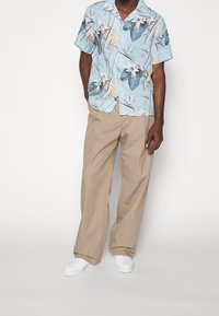 Camicetta azzurro chiaro a maniche corte con stampa di uccelli tropicali, abbinata a pantaloni beige a gamba larga e sneaker bianchi. La camicia presenta un colletto e bottoni.
