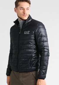 Jachetă puffer neagră cu fermoar frontal, textură matlasată, guler ridicat și buzunare laterale. Prezintă detalii cu logo pe piept.