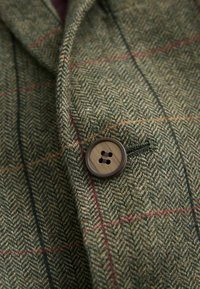 Grön tyg med fiskbensmönster och röda och blå accentfärger. Har en brun knapp med fyra hål som är sydd på slaget.