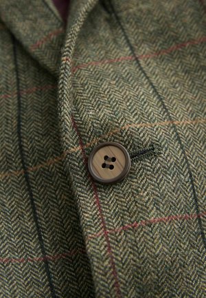Grüner Stoff im Fischgrätenmuster mit roten und blauen Akzenten. Verfügt über einen braunen vierlochn Knopf, der auf dem Revers genäht ist.
