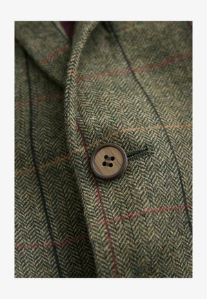 Zelená látka s herringbone vzorom a červenými a modrými akcentmi. Obsahuje hnedý gombík s štyrmi otvormi prišitý na klopu.