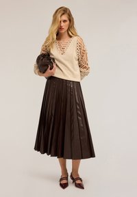 Donna che indossa un maglione a maniche aperte color beige, una gonna midi in pelle plissettata di colore marrone scuro, mentre tiene in mano una borsa a tracolla marrone abbinata.