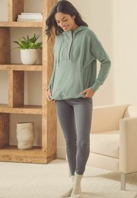 Grøn hættetrøje med afslappet pasform, snørelukning i hætten og kengurulomme, parret med grå ribbede leggings og cremefarvede sokker på en tekstureret gulv.