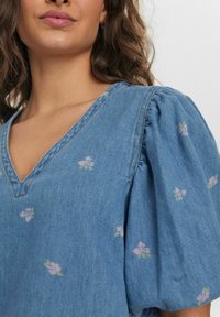 Denimtop med V-ringning och puffärmar; ljusblå med små rosa blommor broderade och kontrasterande sömnadsdetaljer.