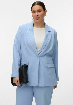 Femeie cu păr închis la culoare, purtând un costum bleu deschis și un top alb, ținând în mână o poșetă neagră, stând în fața unui fundal simplu, deschis la culoare.