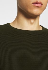 Sweter z okrągłym dekoltem w ciemnozielonym kolorze, z teksturowanym splotem i dopasowanym krojem. Zbliżenie podkreśla gładki dekolt i detale tkaniny.