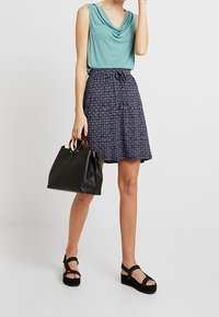Haut drapé vert turquoise clair, jupe à motifs bleu marine, sac à main noir avec poignées marron clair et sandales compensées noires. Tenue décontractée avec une coupe ample.