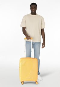 Valise rigide jaune à finition brillante, ornée de motifs ondulés et d'une silhouette de Mickey Mouse, équipée de quatre roulettes et d'une poignée télescopique.