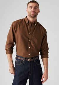 Brown button-up shirt made of cotton with long sleeves, worn untucked over dark denim jeans, accented by a brown belt.