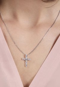 Silver kedja halsband med ett kors hängsmycke prytt med flera klara ädelstenar, placerat mot en ljus rosa tygkrage.