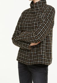 Svart och brun rutig jacka med hög krage, som har ett texturerat tyg och lös passform, kombinerad med mörka jeans.