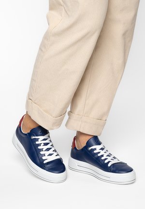 Person, die beige Cordhosen und marineblaue Sneakers mit weißen Sohlen und Schnürsenkeln trägt, steht vor einem weißen Hintergrund.
