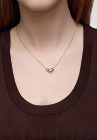 Złota zawieszka w kształcie serca na cienkim łańcuszku, spoczywająca na brązowej bluzce z krótkim rękawem i szerokim dekoltem.