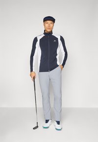 Golf outfit: navy and white zip-up jacket with high collar, gray pants, and white golf shoes. Holding a golf club, wearing a flat cap.