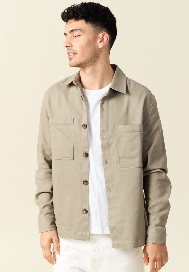 Beige skjorta med knappar i bomull, med krage, två framfickor och bruna knappar, bärs över en vit T-shirt.