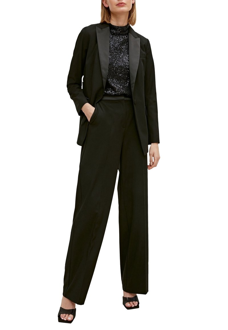 Femme portant un top noir à sequins, un blazer noir avec des revers en satin, un pantalon noir à jambes larges et des talons ouverts noirs.