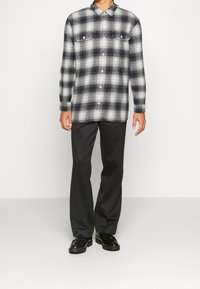 Chemise en flanelle à carreaux noir et blanc avec deux poches poitrine, associée à un pantalon gris foncé et des chaussures noires brillantes. Tissu texturé, col chemise.