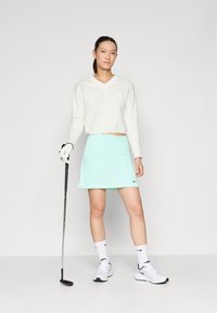 Λευκή μακριά μανίκια μπλούζα v-neck, ανοιχτού μέντας φούστα και λευκά αθλητικά Nike με μαύρες λεπτομέρειες. Κράτησε μια γκολφ κλαμπ στο δεξί χέρι, στέκεται σε απλό φόντο.