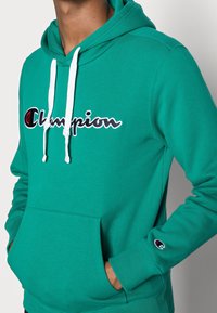 Sinivihreä huppari, jossa valkoiset nauhat ja etutasku. Musta-punaisella kirjotulla "Champion"-logolla varustettu. Pehmeää fleecemateriaalia.