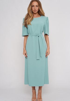 Kobieta z długimi blond włosami, ubrana w jasnoniebieską sukienkę midi z bufkami do łokcia i wiązaniem w talii, stojąca na jednolitym tle.