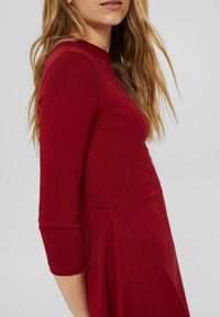 Rotes Kleid mit enganliegendem Oberteil, Rundhalsausschnitt und Dreiviertelärmeln. Das Material erscheint weich und dehnbar, mit einer glatten Textur.