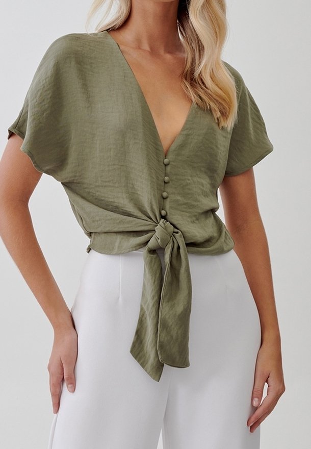 Femme portant un chemisier vert olive à manches courtes avec des boutons devant et un nœud noué, associé à un pantalon blanc taille haute.