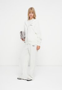 Bijela zip-up hoodie i odgovarajuće široke trenirke. Hoodie ima prednji džep i izvezene detalje. Mekana tekstura.