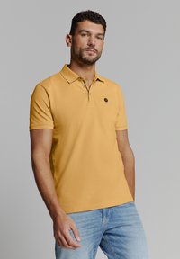 Gele polo met korte mouwen, een kraag en een klein zwart embleem op de borst. Gecombineerd met lichtblauwe denim jeans.